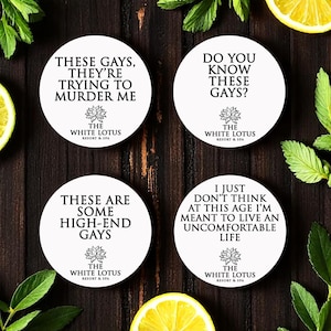 Puede incluir: Cuatro posavasos redondos blancos con texto negro y un logotipo de flor de loto. Los posavasos tienen frases como "These gays, they're trying to murder me". Están sobre una superficie de madera oscura.