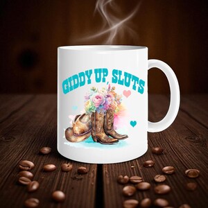 Op de afbeelding: Witte keramische mok met de tekst "GIDDY UP, SLUTS" in turquoise. De mok heeft een aquarelontwerp van cowboylaarzen, een hoed en een boeket bloemen. Twee harten en een turquoise hart zijn ook in het ontwerp opgenomen.