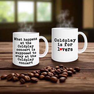 May include: Two white ceramic mugs with black text on a wooden surface. One mug reads "What happens at the Coldplay concert is supposed to stay at the Coldplay concert." The other mug says "Coldplay is for lovers" with a red heart.