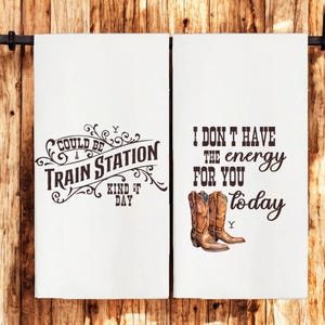 Może przedstawiać: Dwa białe ręczniki kuchenne z brązowym tekstem i obrazkami. Lewy ręcznik mówi "Could be a train station kind of day". Prawy ręcznik mówi "I don't have the energy for you today" i przedstawia parę brązowych kowbojskich butów.