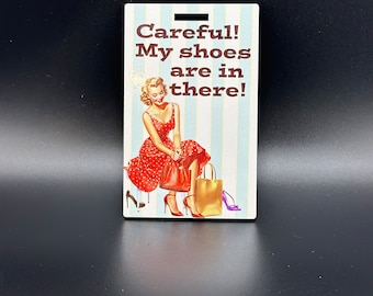 May include: A vintage-style sign with a woman in a red dress and white polka dots sitting on a bench with a shopping bag. The sign reads "Careful! My shoes are in there!"