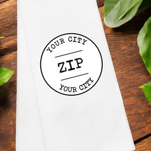 以下が含まれることがあります： 「YOUR CITY ZIP YOUR CITY」と書いてある黒い円形のデザインが施された白いキッチンタオルです。