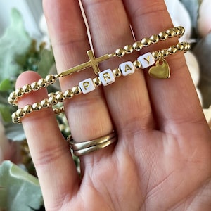 May include: Two gold beaded bracelets. One features a gold cross and the word "PRAY" spelled out in white letter beads. The other bracelet has a gold heart charm. The bracelets are worn on a wrist with gold rings.