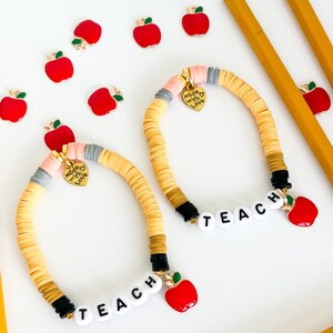 May include: Two bracelets with yellow, pink, and gray beads, black beads, and white letter beads spelling "TEACH". Each bracelet has a small gold charm with the words "Made with Love". Each bracelet also has a red apple charm.