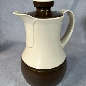 May include: A two-tone coffee carafe featuring a cream-colored upper body and a dark brown lower section. The vessel includes a matching dark brown lid with a central knob, a curved handle, and a wide pouring spout, creating a sleek silhouette.