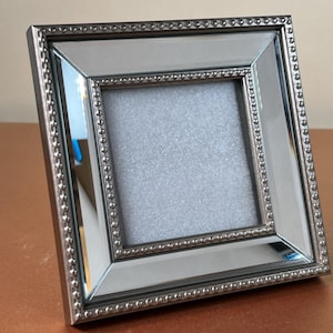 May include: A square picture frame featuring a reflective mirrored border and a metallic silver beaded trim. The centre of the frame contains a textured, light grey square insert. The frame has a bevelled design that adds depth to the structure.