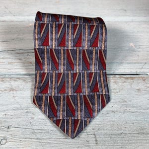 May include: A necktie featuring a repeating geometric pattern of rectangular blocks. Each block contains a diagonal stripe in deep red and slate blue, separated by thin vertical lines in a muted tan color against a textured grey background.