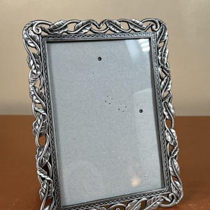 May include: A rectangular picture frame with a metallic silver finish. The border features a raised, repeating leaf and vine motif. The inner edge of the frame has a fine beaded texture surrounding a plain, light gray rectangular insert.