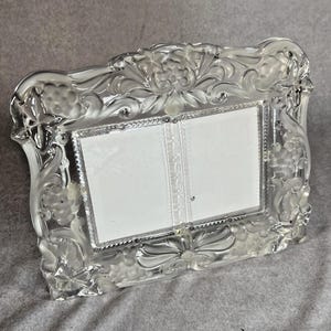 May include: A clear glass picture frame featuring a rectangular center opening and an ornate border decorated with raised, frosted relief carvings of grape clusters, leaves, and scrolling vine patterns throughout the entire frame edge.