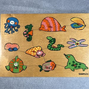 May include: A rectangular wooden peg puzzle featuring colorful illustrations of various sea creatures including an octopus, seahorse, fish, clam, and seal. Each piece has a small red knob for handling. The word SIMPLEX is printed in the bottom right corner.