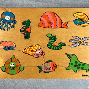 Può includere: Puzzle in legno con creature marine colorate, tra cui un polpo, un cavalluccio marino e pesci. I pezzi del puzzle sono collegati da linee e la parola "SIMPLEX" è stampata in basso a destra.