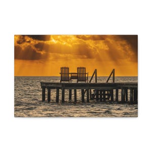 May include: A scenic photograph of a wooden pier extending into the ocean at sunset. Two wooden chairs and a small table are on the pier. The sky is filled with golden light and dramatic clouds.