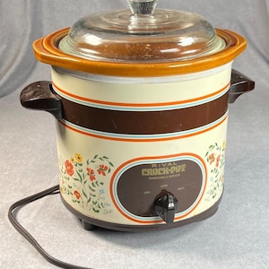 May include: Vintage Rival Crock-Pot with a glass lid. The slow cooker has a cream-colored body with brown and orange accents, floral patterns, and a control dial. The power cord is plugged in.