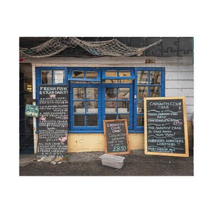 May include: A jigsaw puzzle featuring a seaside shop with blue window frames and multiple signs. The signs advertise fresh fish and crab daily, sandwiches, and crab sandwiches. A fishing net hangs above the shop.