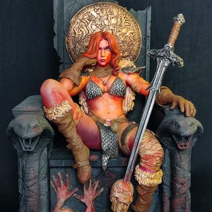 May include: A detailed figurine of a warrior woman with red hair, seated on a throne. She wears a metal bikini, boots, and holds a sword. The throne is adorned with snakes, skulls, and a golden shield.