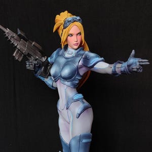 Peut inclure: Une figurine en résine peinte représentant un personnage blond en armure futuriste bleue et blanche, tenant un long fusil. Le personnage se tient sur une base rouge charnue et texturée, incrustée de plusieurs crânes et de pointes osseuses.