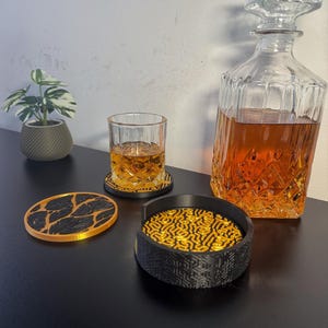 Op de afbeelding: Een heldere glazen karaf gevuld met amberkleurige vloeistof, een whiskyglas met amberkleurige vloeistof en een set decoratieve onderzetters. De onderzetters hebben een zwart-goud design. Een kleine potplant is ook zichtbaar.
