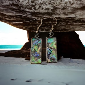May include: A pair of silver earrings with iridescent green and blue abalone shell rectangular pendants. The earrings are photographed on a white sandy beach with a large rock in the background.