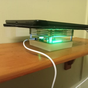 May include: A white charging stand with a clear acrylic frame and a green LED light. A black tablet is resting on top of the stand.