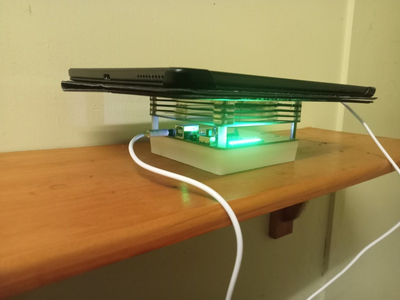 May include: A white charging stand with a clear acrylic frame and a green LED light. A black tablet is resting on top of the stand.