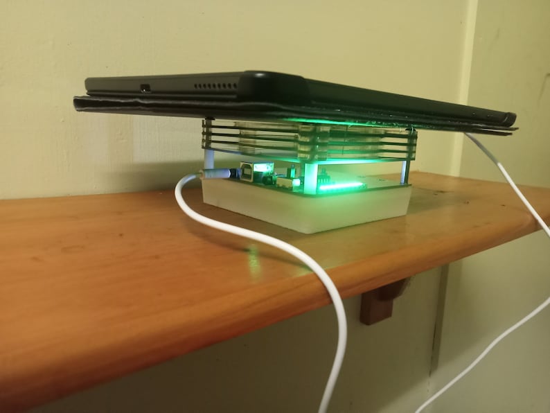 May include: A white charging stand with a clear plastic case and a green light. A black tablet is resting on top of the stand. A white charging cable is plugged into the stand.