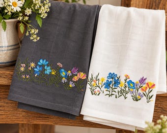 May include: Two rectangular kitchen towels, one dark charcoal grey and one white, featuring a bottom border of embroidered wildflowers in shades of blue, yellow, pink, and purple with green stems and leaves, crafted from a textured fabric.