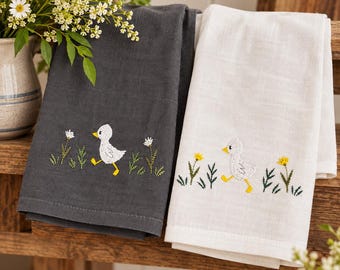 Könnte beinhalten: Zwei Küchentücher, eines dunkelgrau und eines weiß, mit gestickten Motiven eines kleinen weißen Entleinkükens, das zwischen grünem Gras und gelben oder weißen Blumen läuft. Die Tücher liegen gefaltet auf einer Holzoberfläche neben einer Vase.