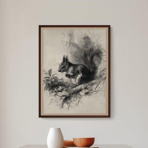 Könnte beinhalten: Ein gerahmter Wandkunstdruck im Kohle-Stil mit einer detaillierten Illustration eines Eichhörnchens, das auf einem Ast inmitten von Laub sitzt. Das Kunstwerk ist in Schwarz-, Grau- und Cremetönen gehalten und in einem schlichten dunkelbraunen Rahmen.