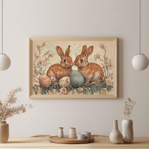May include: A rectangular wall art print in a light wood frame featuring two brown rabbits sitting among light-colored flowers and three patterned Easter eggs. The artwork is displayed on a plain wall above a wooden dining table with vases.