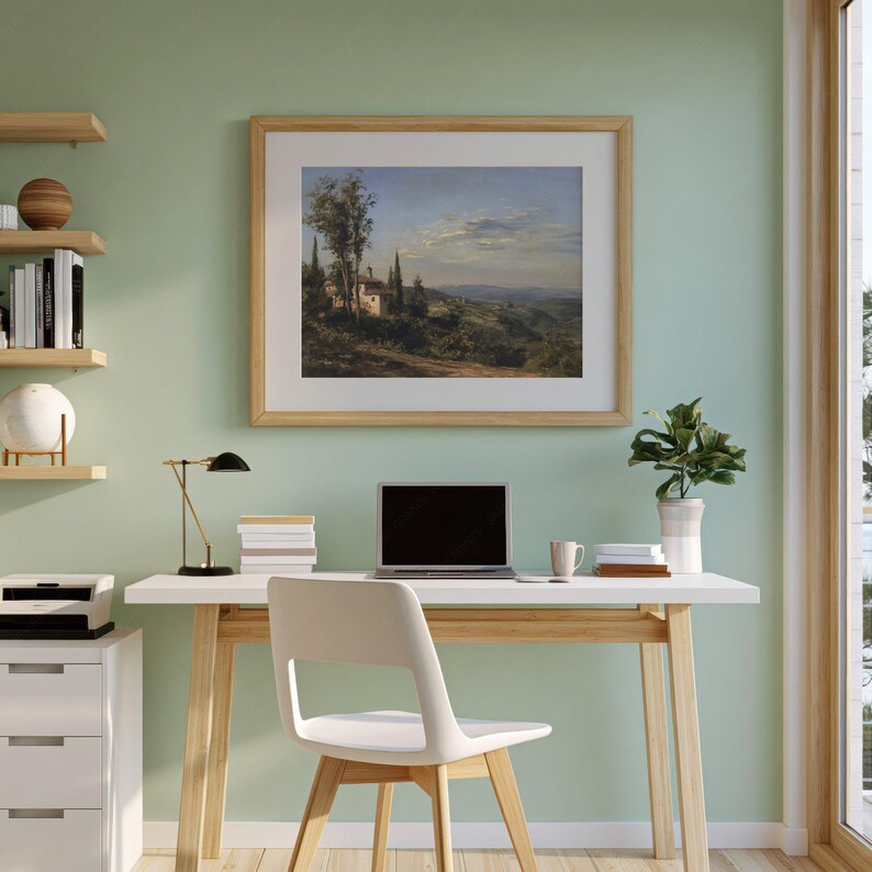 May include: A framed landscape painting depicting a rural scene with buildings, trees, and rolling hills. The artwork is displayed above a white desk with a laptop, lamp, and chair. The room has light green walls and wooden shelves.