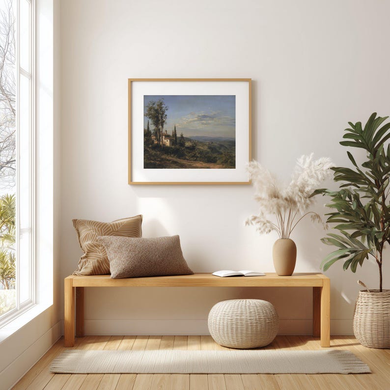 May include: A framed landscape painting depicting a Tuscan villa scene with rolling hills, trees, and a blue sky. The artwork is displayed above a wooden bench with decorative pillows, a woven ottoman, and a potted plant, creating a cozy interior design.