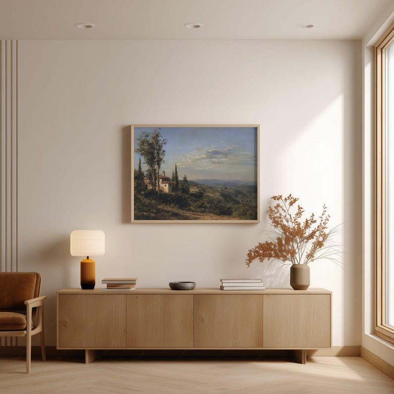 May include: A framed landscape painting depicts a Tuscan villa scene with rolling hills, trees, and a cloudy sky. The artwork hangs above a wooden cabinet with a lamp, books, and a vase with dried flowers.
