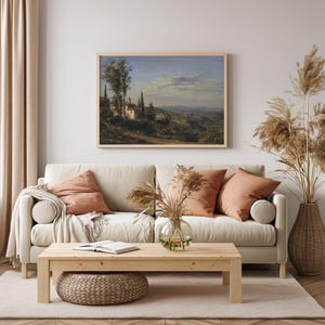May include: A framed landscape painting depicts a Tuscan villa scene with rolling hills, trees, and a cloudy sky. The painting hangs above a beige sofa with peach-colored pillows. A wooden coffee table and woven ottoman complete the living room.
