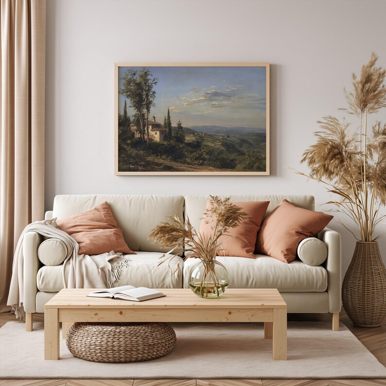 May include: A framed landscape painting depicts a Tuscan villa scene with rolling hills, trees, and a cloudy sky. The painting hangs above a beige sofa with peach-colored pillows. A wooden coffee table and woven ottoman complete the living room.