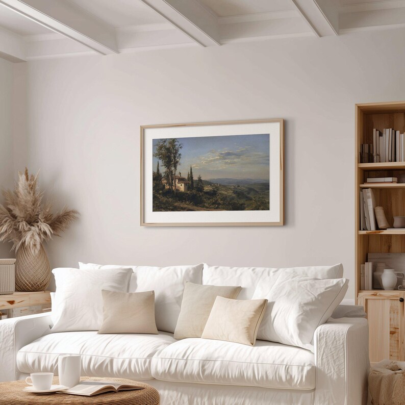 May include: A framed landscape painting depicting a European village scene with rolling hills, trees, and a cloudy sky. The artwork is displayed above a white sofa with beige pillows, enhancing the room's decor.