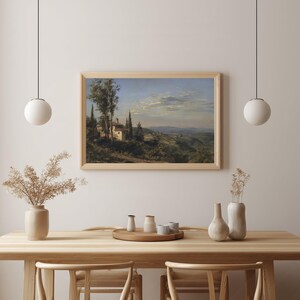 May include: A framed landscape painting depicts a Tuscan villa nestled among cypress trees and rolling hills under a cloudy sky. The artwork is displayed above a wooden table with decorative vases and a tray.