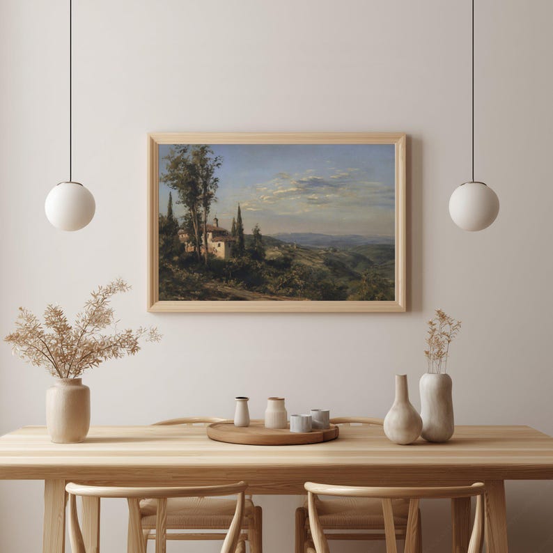 May include: A framed landscape painting depicts a Tuscan villa nestled among cypress trees and rolling hills under a cloudy sky. The artwork is displayed above a wooden table with decorative vases and a tray.