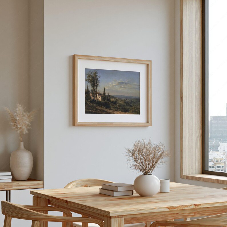 May include: A framed landscape painting depicting a European village scene with a light brown wooden frame. The artwork features a village with trees and a blue sky. The painting is displayed on a light-colored wall in a room with a wooden table and chairs.