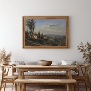 May include: A framed landscape painting depicts a Tuscan scene with a white building, trees, and rolling hills under a blue sky. The artwork is displayed in a natural wood frame, adding a touch of rustic elegance to the room.