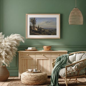 May include: A framed landscape painting hangs above a wooden cabinet. The painting depicts a rural scene with buildings and trees. A woven pendant light and a wicker chair with a green blanket are also visible. A large vase of pampas grass sits to the left.