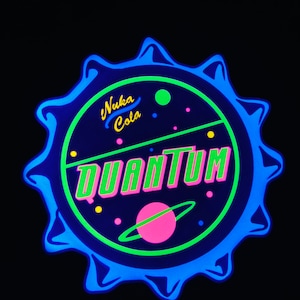 May include: A neon blue and green Nuka Cola Quantum bottle cap design. The design features the words "Nuka Cola" in yellow and "Quantum" in green and pink, with a pink planet and green and yellow dots.