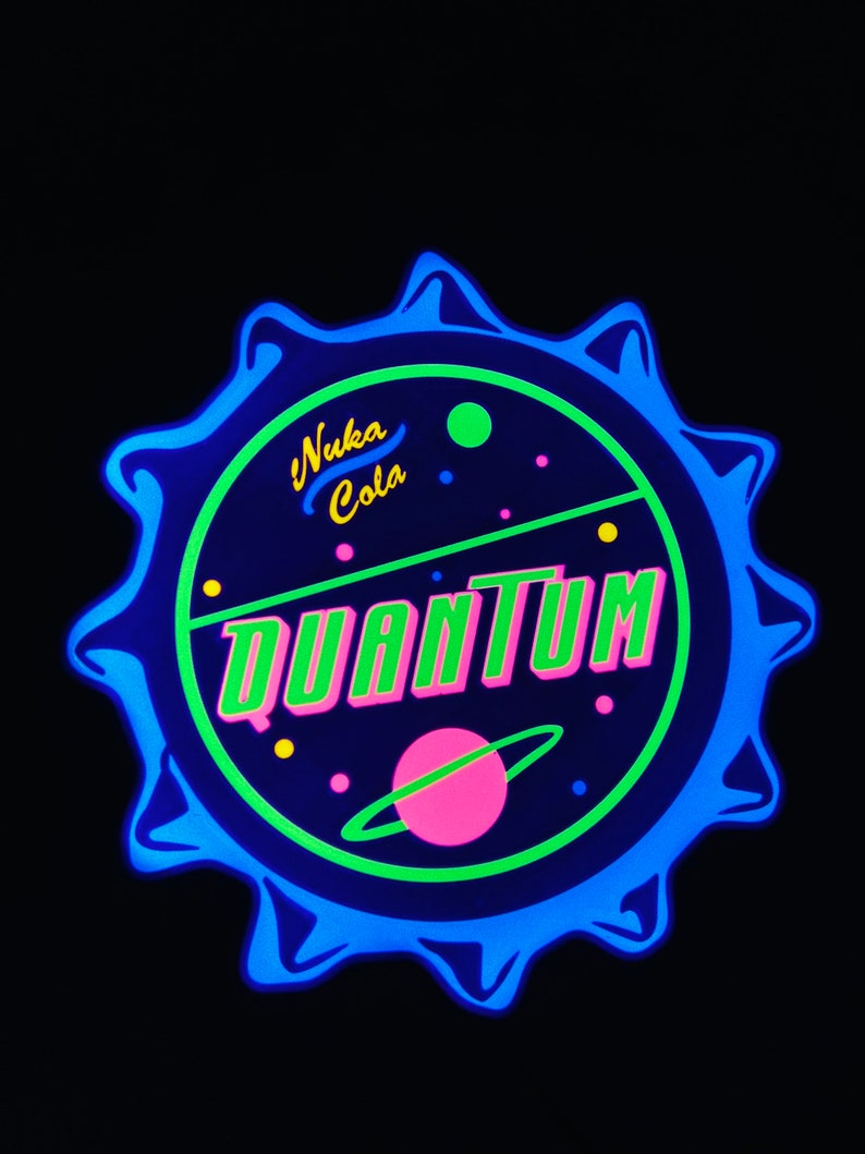 May include: A neon blue and green Nuka Cola Quantum bottle cap design. The design features the words "Nuka Cola" in yellow and "Quantum" in green and pink, with a pink planet and green and yellow dots.