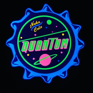 May include: A neon sign in the shape of a bottle cap, featuring the words "Nuka Cola Quantum" in green and pink. The design includes a planet with a ring and small dots, all against a dark blue background.