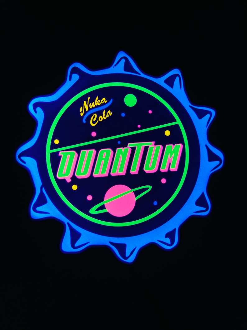 May include: A neon sign in the shape of a bottle cap, featuring the words "Nuka Cola Quantum" in green and pink. The design includes a planet with a ring and small dots, all against a dark blue background.