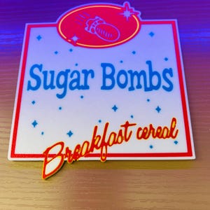 May include: A rectangular sign with a red border and the words "Sugar Bombs" in blue, and "Breakfast cereal" in red script. A red oval with a bomb graphic is at the top. Blue diamond shapes are scattered across the white background.