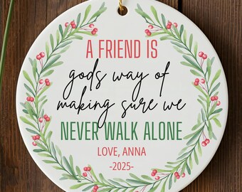 May include: A white ceramic ornament with a gold string for hanging. The ornament features the text "A FRIEND IS gods way of making sure we NEVER WALK ALONE" in red and black script, surrounded by a green and red floral wreath. The text "LOVE, ANNA -2025-" is also included.