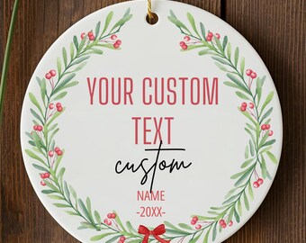 May include: A round, white ceramic ornament with a gold string for hanging. The ornament features a watercolor wreath of green leaves and red berries. Text in red and black reads "YOUR CUSTOM TEXT custom NAME -20XX-" with a red bow at the bottom.