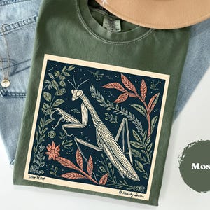 May include: A moss green t-shirt featuring a detailed graphic of a praying mantis surrounded by foliage in a dark blue square. The design includes orange and green leaves and flowers. The shirt is laid on top of blue jeans and a tan hat.