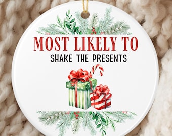 May include: A white, round Christmas ornament with the text "MOST LIKELY TO SHAKE THE PRESENTS" in red. The ornament features illustrations of wrapped gifts with red bows and a candy cane, surrounded by green pine branches.