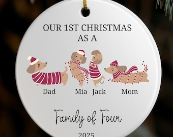 May include: White ceramic Christmas ornament with a gold string. The ornament features a dachshund family illustration with the text "Our 1st Christmas as a Family of Four 2025". The dachshunds are wearing festive attire.